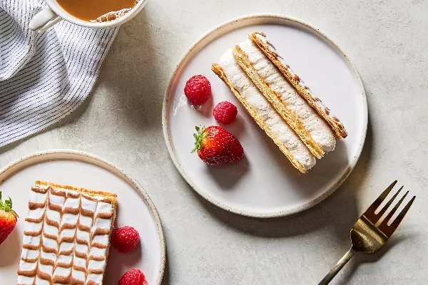  Deux morceaux de gâteau Mille-Feuille sur des assiettes, présentés avec des baies, une tasse de café et une fourchette dorée.