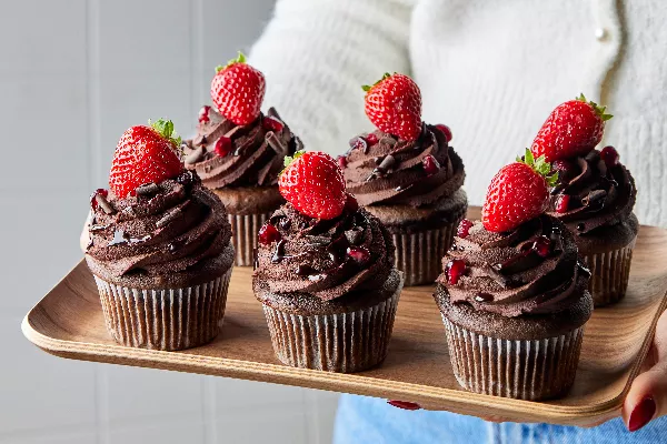 Six cupcakes au chocolat et au vin rouge garnis de ganache, de copeaux de chocolat, de grenade et de fraises sur un plateau en bois tenu par une femme en pull blanc et en jean