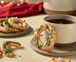 Des biscuits en sucre glacé avec du glaçage et décorés comme des couronnes avec du romarin, l'un sur une soucoupe avec une tasse de café, un autre cassé sur une table, et plusieurs sur une assiette, présentés dans un cadre festif.