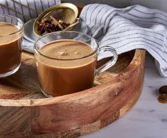 Deux tasses en verre de masala chai sur un plateau de service rond en bois, accompagnées d'une passoire dorée contenant des épices entières, d'un torchon et d'une cuillère dorée.