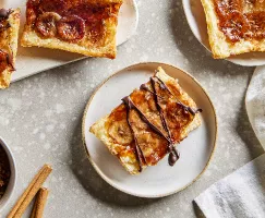 Quatre tartes renversées à la banane, deux dans des assiettes et deux sur un plateau, présentées avec un bol de sucre brun et des bâtonnets de cannelle.