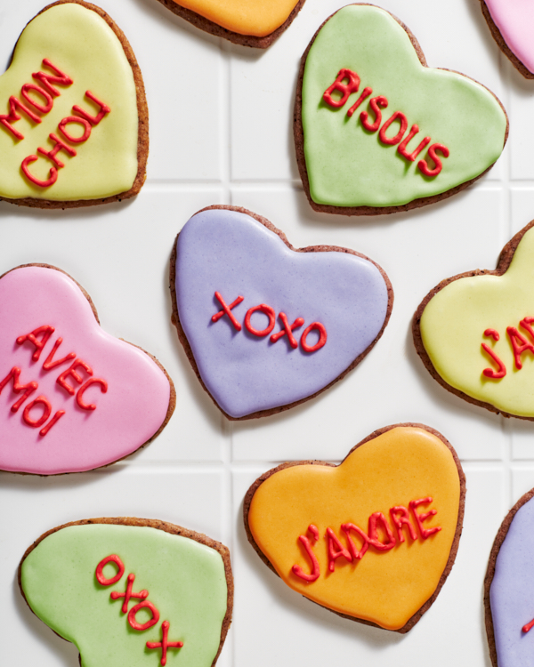 Biscuits en sucre en forme de cœur décorés avec du glaçage royal aux couleurs pastel avec des messages romantiques écrits dans le glaçage, présentés sur des carreaux blancs.