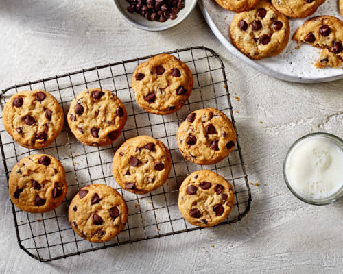 Des biscuits aux pépites de chocolat sur une grille de refroidissement en fil métallique, avec des biscuits supplémentaires sur un plateau, montrés avec un bol de pépites de chocolat et un verre de lait blanc. 