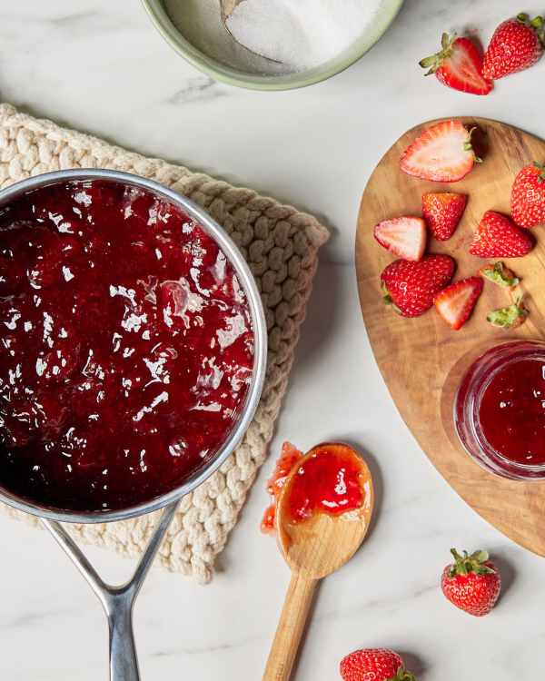 Un pot de confiture de fraises fraîche sur un torchon, présenté avec des fraises tranchées, une cuillère en bois et un bol de sucre granulé.