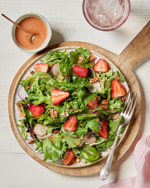Une salade verte dans une assiette posée sur une planche de service en bois, garnie de tranches de fraises, présentée avec un bol de vinaigrette aux fraises.