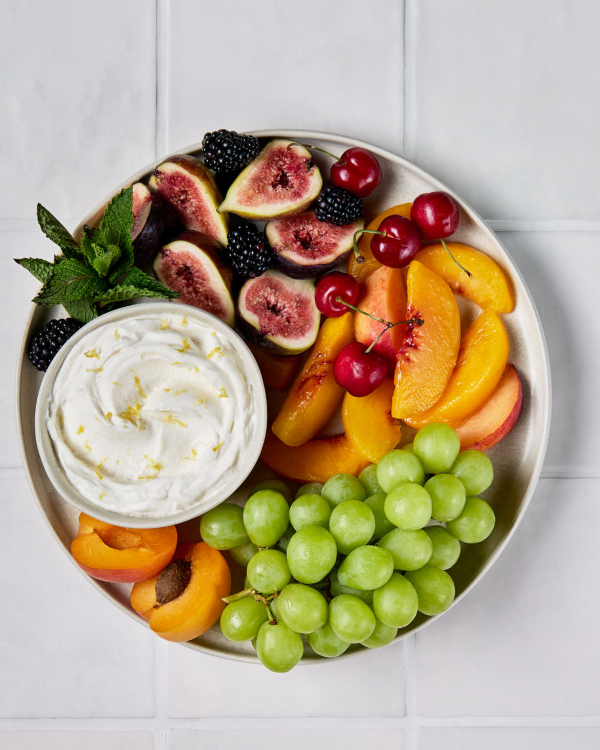 Un plateau de fruits est présenté sur un comptoir carrelé, avec un bol de crème fouettée au citron, des figues, des mûres, des pêches, des cerises et des raisins.