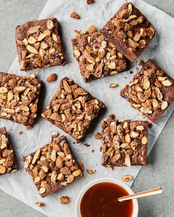  Des carrés de brownies aux noix sucrées et épicées garnies de caramel sur un morceau de papier ciré déposé sur un comptoir de cuisine avec un bol de caramel.
