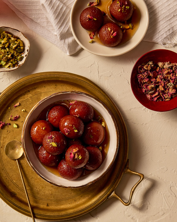 Des gulab jamuns arrosés de sirop et garnis de morceaux de pistaches et de pétales de rose, répartis entre deux assiettes, et présentés avec des fourchettes en or.