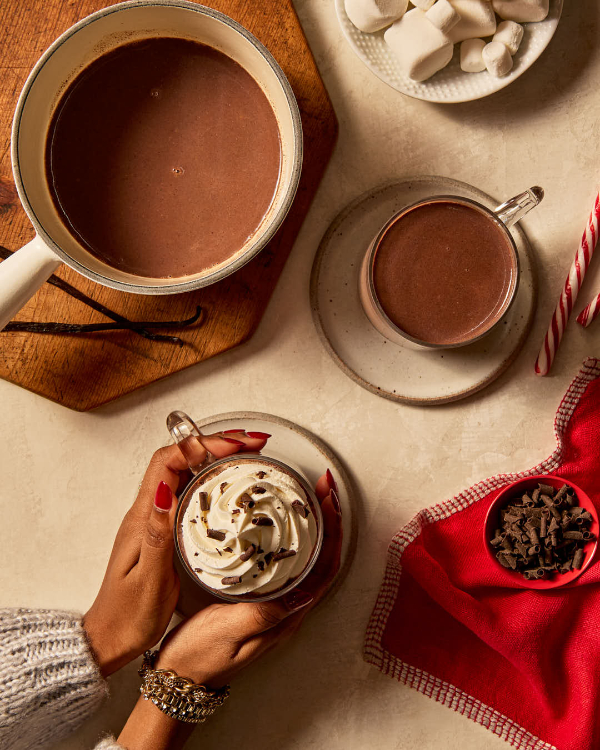 Deux mains féminines tenant une tasse en verre de chocolat chaud à la vanille, garnie de crème fouettée et agrémentée de chocolat râpé, reposant sur une soucoupe et accompagnée de guimauves, de chocolat râpé, de bâtons de bonbon et d'une autre tasse de chocolat chaud.