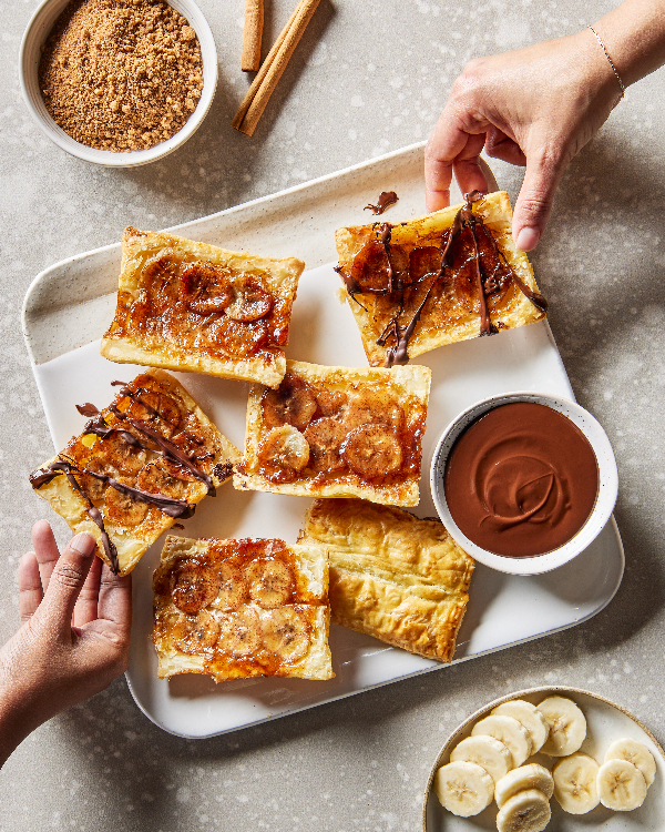 Deux mains atteignant des tartes renversées à la banane sur un plat blanc, présentées avec un bol de sauce au chocolat, de sucre brun, de bâtonnets de cannelle et de bananes tranchées.