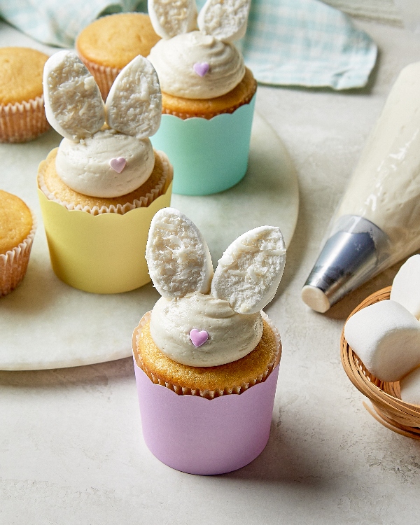 Cupcakes au citron décoratifs sur le thème de Pâques avec un glaçage à la crème au beurre et des oreilles de lapin en guimauve trempées dans la noix de coco, présentés dans des caissettes à cupcakes pastel, parfaits pour un dessert de printemps festif.