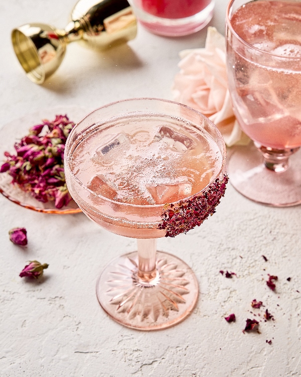 Deux verres à tige uniques de spritz litchi et rose sur glaçons. Des pétales de rose séchés garnissent les verres qui sont présentés dans un cadre neutre avec une rose fraîche en arrière-plan, une assiette de pétales de rose séchés et un doseur doré.