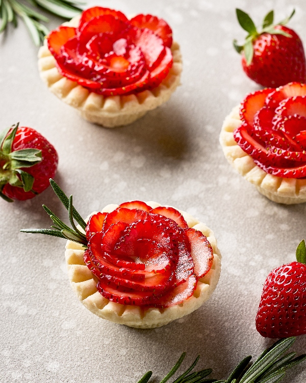  Trois mini-tartelettes fraises et romarin sur un comptoir de cuisine avec trois fraises fraîches et quelques brins de romarin.