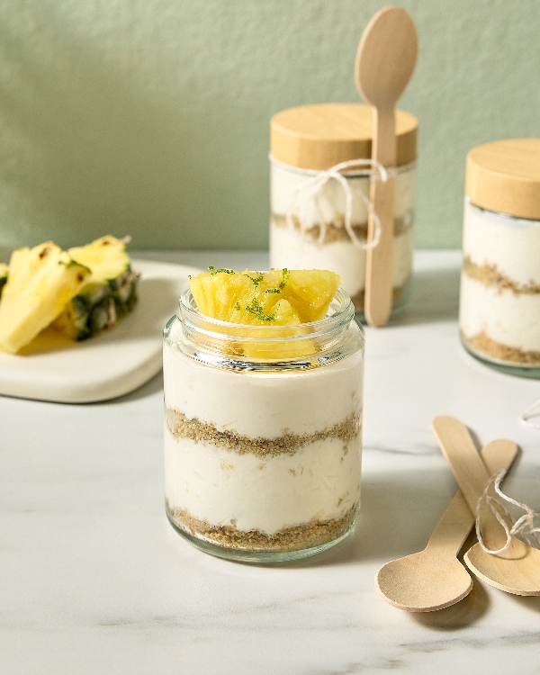 Plusieurs verrines de cheesecake à l'ananas en portions individuelles sont disposées sur une surface en marbre blanc. Chaque verrine présente des étages de gâteau au fromage crémeux et de croûte friable au graham cracker, garnies de morceaux d'ananas frais et d'une pincée de zeste de lime. Des cuillères en bois attachées avec de la ficelle agrémentent les verrines, et une assiette débordant de morceaux d'ananas supplémentaires est visible en arrière-plan.