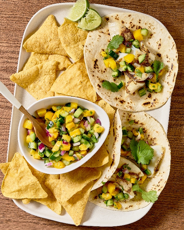 Sur une table en bois, une assiette blanche présente deux tacos au poisson garnis d'une salsa à la mangue et au concombre, le tout rehaussé de coriandre fraîche. À côté des tacos, un bol de salsa, avec une cuillère en bois, est entouré de chips tortillas bien croustillants.