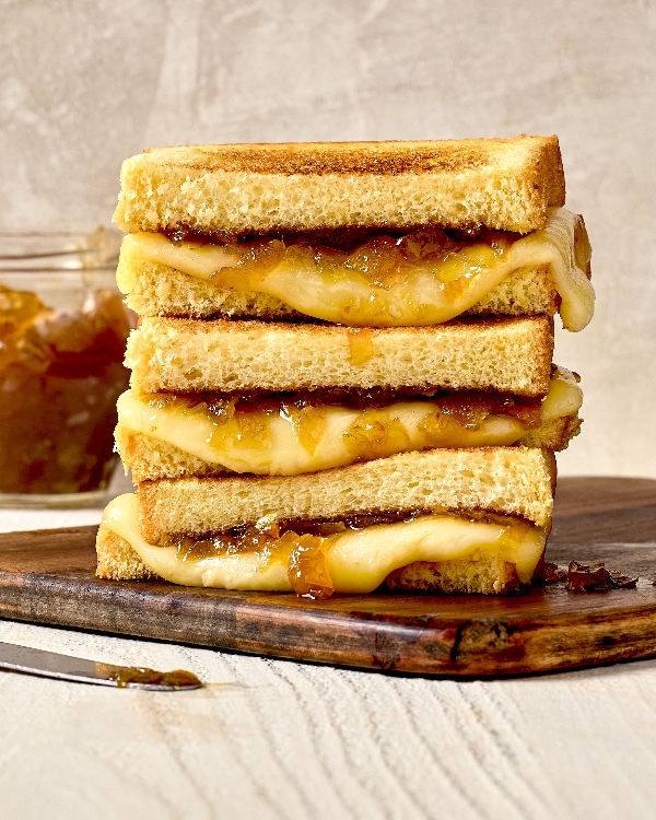 Une pile de demi-sandwichs au fromage grillé à la confiture d’oignons disposée sur une planche en bois. Le pain des sandwichs est doré, le fromage est fondant et la confiture d’oignons caramélisés déborde légèrement. En arrière-plan, un pot de confiture d’oignons et un couteau avec de la confiture sont partiellement visibles.