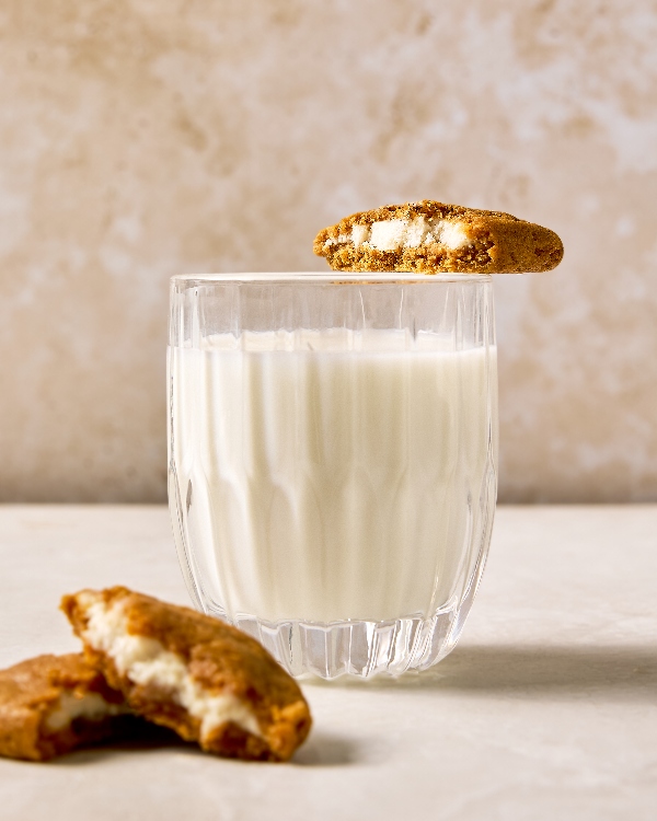 Gros plan d’un verre de lait avec un biscuit aux épices pour tarte à la citrouille farci au fromage à la crème reposant sur le bord. Le biscuit est cassé en deux, révélant la garniture au fromage à la crème. Un autre biscuit est placé devant le verre, mettant en évidence la garniture riche et crémeuse à l’intérieur du biscuit doré