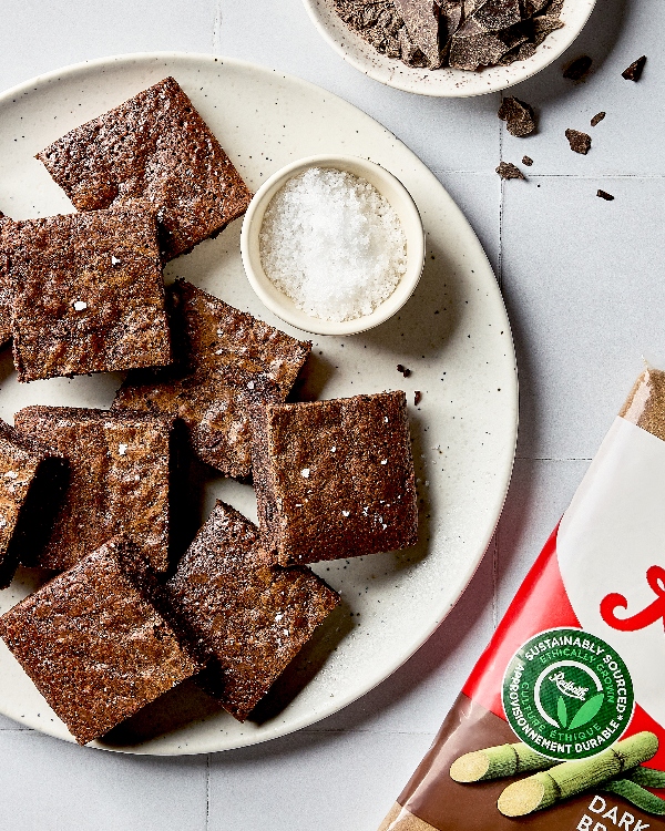 Une assiette de brownies garnis de sel en flocons présentée sur un comptoir en carreaux blancs avec un bol de sel en flocons, un bol de chocolat noir haché et un sac de cassonade.