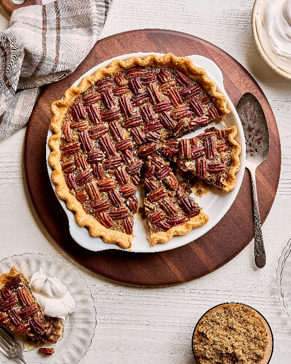 Une tarte au pacane dans un plat à gratin, montrant une tranche en train d'être servie sur un léve-tarte par une personne portant un tablier, sur une table en bois blanc avec un bol de pacanes.