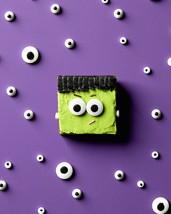 Un brownie au chocolat décoré de glaçage et de bonbons pour ressembler au monstre de Frankenstein, présenté sur un fond violet avec d'autres bonbons pour les yeux éparpillés autour.