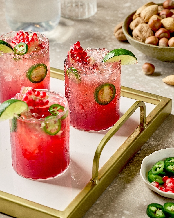 Trois verres de Fizz Épicé à la Grenade et au Gingembre disposés sur un plateau à bordure dorée. Chaque boisson est garnie de quartiers de lime, de graines de grenade et de tranches de jalapeño frais. Les cocktails rouges vifs se détachent sur le fond blanc du plateau, créant une présentation élégante. À l'arrière-plan, on trouve un bol de noix mélangées et un petit plat avec des tranches de jalapeño et des graines de grenade supplémentaires.