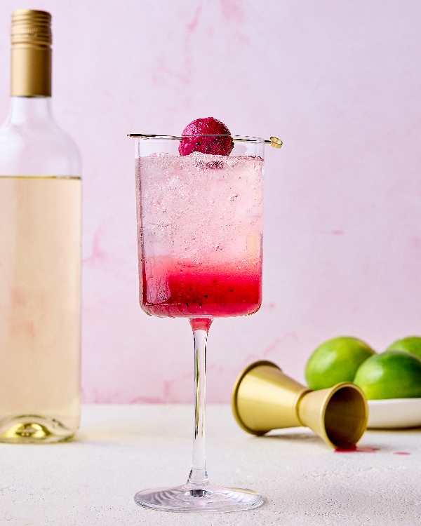 Un verre de spritz à la pitayaha servi sur glace, avec un dégradé de rose et garni d’une boule de pitayaha sur brochette. En arrière-plan, une bouteille de vin blanc, un doseur à cocktail doré et des limes sont présentés sur un fond rose pâle.