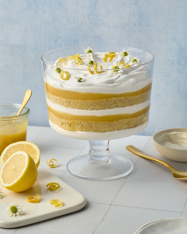 Un bol à bagatelle en verre rempli d’une bagatelle au citron et à la fleur de sureau avec ses couches de gâteau éponge, de curd au citron et de crème fouettée. Le dessert est couronné de rubans de zeste de citron et de petites fleurs blanches. Des citrons frais, un pot de curd au citron et une cuillère dorée sont dispersés autour du bol de service, le tout sur un fond bleu doux.