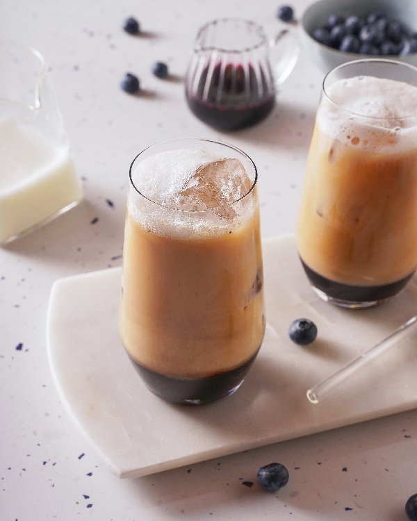 Des cafés glacés servis dans de grands verres avec des couches visibles de sirop de bleuets, de café et de lait. Les boissons sont garnies de gros glaçons et placées sur une planche de service en pierre blanche. À l'arrière-plan, il y a un petit pot de lait, un pichet en verre de sirop de bleuets, un bol de bleuets frais et des bleuets éparpillés sur un comptoir moucheté.