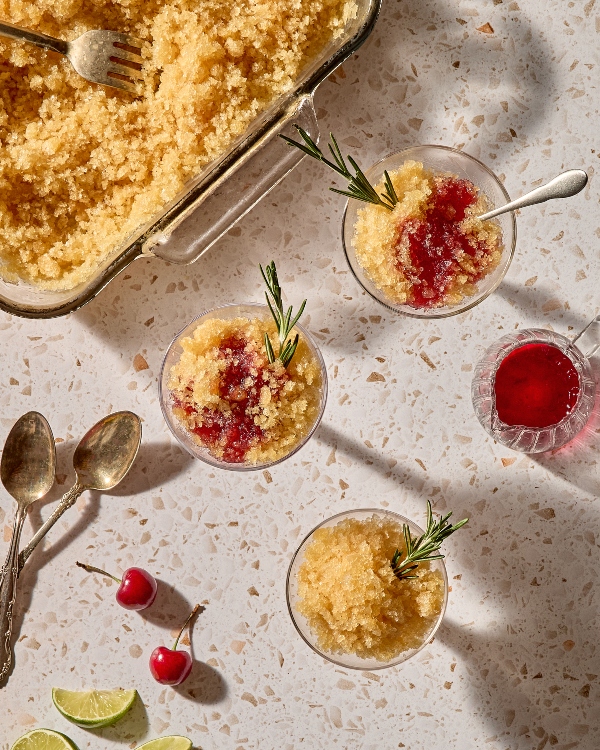  Vue du dessus de trois tasses en verre remplies d’un dessert glacé doré, certains nappés d’un sirop rouge profond et garnis de brins de romarin. À proximité se trouvent des cuillères rétro, des cerises fraîches, des quartiers de lime et une carafe en verre contenant du sirop de cerise rouge. Un grand plat en verre contenant la slush dorée avec une fourchette est placé dans le coin supérieur gauche sur une surface en terrazzo moucheté.