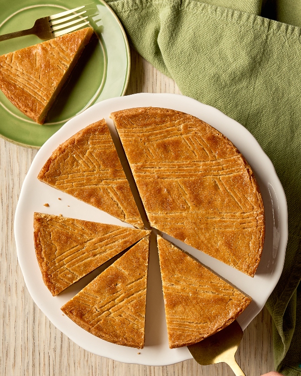 Un plan rapproché en plongée d'un gâteau au beurre hollandais (Boterkoek) rond, doré et découpé en six pointes, disposé sur une assiette de service blanche. Le gâteau présente une texture dense et beurrée et est orné d'un motif quadrillé en losanges sur toute sa surface. Une main utilisant une spatule de service dorée retire une pointe par le bas à droite. En haut à gauche, une pointe est déjà déposée sur une petite assiette verte à côté d'une fourchette, avec une serviette verte froissée à proximité.