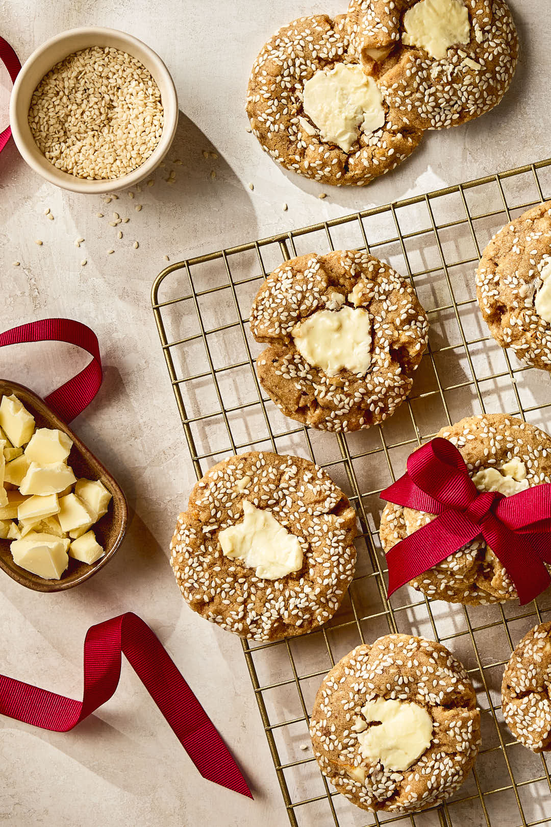 Des biscuits au tahini et aux morceaux de chocolat blanc, enrobés de graines de sésame, refroidissent sur une grille; l'un d'eux est attaché avec un ruban rouge. Un bol de graines de sésame, un plat de chocolat blanc haché et un ruban rouge dénoué sont disposés autour des biscuits sur une surface texturée pâle.