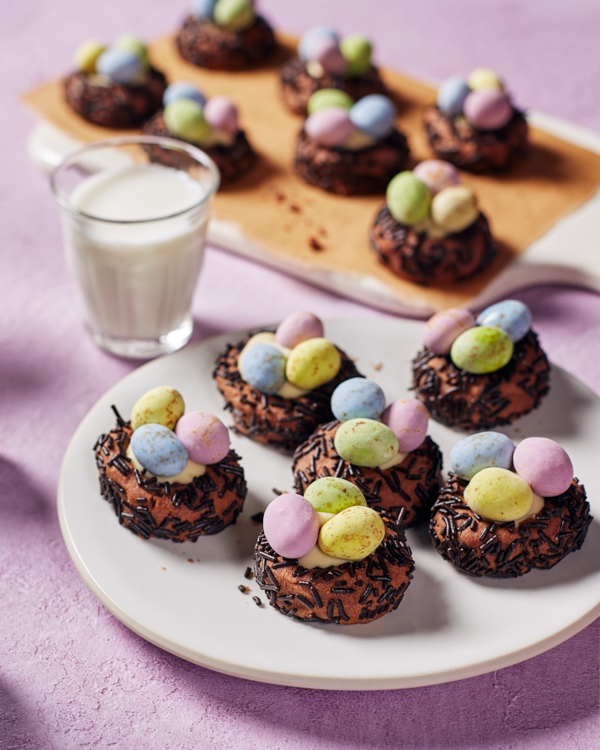 Biscuits chocolatés en forme de nids d'oiseaux, garnis de chocolat blanc et d'œufs en chocolat enrobés de bonbons, sur une assiette et une planche à découper.