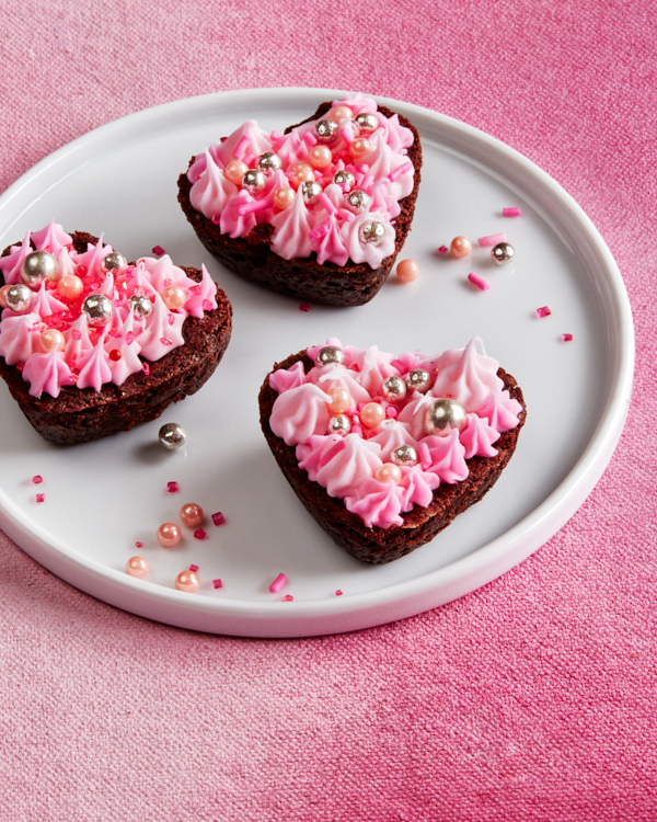 Trois bouchées de brownies double-chocolat recouvertes de glaçage rose et de décorations dans une assiette sur une nappe rose.