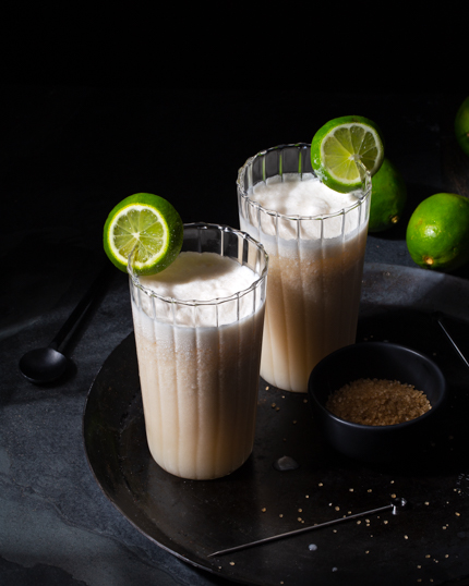 Deux verres de kombucha à la noix de coco garnis de tranches de lime, sur une assiette noire avec des limes et un bol de Sucre Turbinado Simplement Brut™