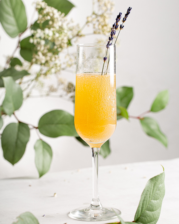 Un grand verre de mimosa citron et lavande décoré de brins de lavande fraîche avec de la verdure et des fleurs en arrière-plan>