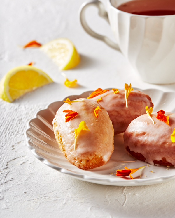 Des madeleines au citron glacées et décorées de pétales de fleurs comestibles, disposées sur une soucoupe et accompagnées d’une tasse de thé.
