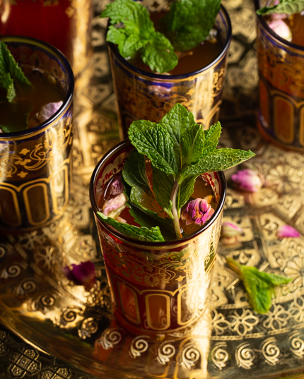 Cinq verres colorés remplis de thé marocain à la menthe, décorés de feuilles de menthe et disposés sur un plateau doré