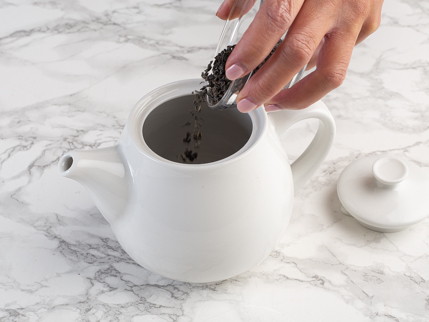 Feuilles de thé vert qui se font transférer d’un bol en verre dans une théière blanche