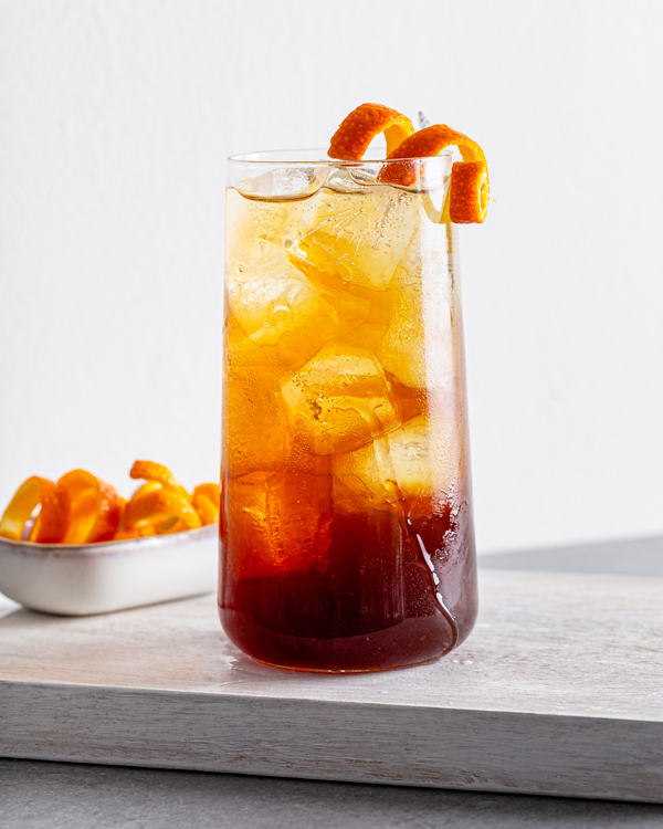 Un verre de spritz café infusé à froid et orange avec glaçons et zeste d’orange, et une assiette de bandelettes frisées de zeste d’orange.