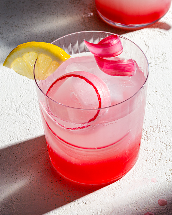 Verre de limonade à la rhubarbe rose garni d'une boucle de rhubarbe, posé sur un comptoir avec le soleil qui brille à travers une fenêtre.
