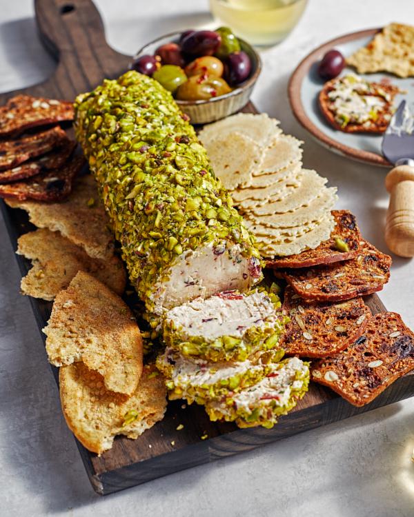 Une bûche de fromage à la crème enrobée de pistaches concassées sur une planche de service accompagnée de craquelins, de croustilles et d’un bol d’olives