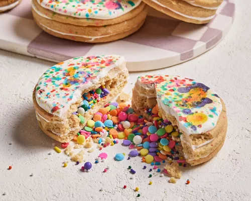 Un biscuit en forme de piñata glacé, brisé en deux avec des confettis are-en-ciel et des vermicelles sortis, présenté avec trois autres biscuits sur un plateau à carreaux blancs et mauves en arrière-plan