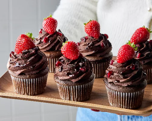 Six cupcakes au chocolat et au vin rouge garnis de ganache, de copeaux de chocolat, de grenade et de fraises sur un plateau en bois tenu par une femme en pull blanc et en jean.