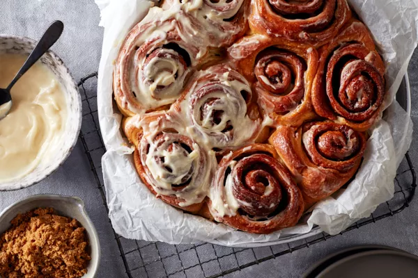 Un plat de cuisson rond tapissé de papier sulfurisé et rempli de petits pains à la cannelle, dont la moitié est glacée, présenté avec un bol de glaçage et un bol de Cassonade Foncée.