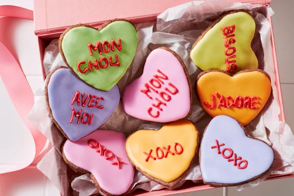 Des biscuits en sucre en forme de cœur décorés de glaçage royal avec des messages romantiques écrits dans le glaçage, présentés sur des carreaux blancs avec des bols de glaçage royal et une poche à douille.
