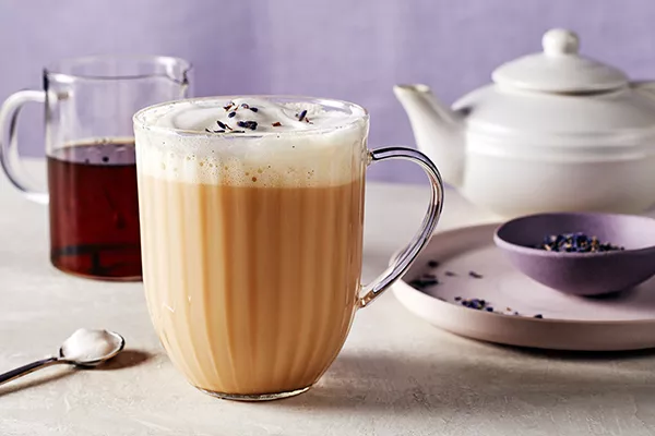 Un verre à latte London Fog avec mousse de lait, montré avec une carafe de sirop de vanille, une théière, une cuillère à mélanger et un bol pourpre de thé Earl Grey en vrac sur un plateau.