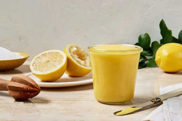 Un pot en verre rempli de lemon curd est présenté avec un citron coupé en deux, dont une moitié est pressée, un presse-citron et un bol de sucre granulé.