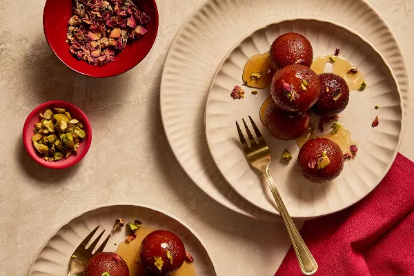  Des gulab jamuns arrosés de sirop et garnis de morceaux de pistaches et de pétales de rose, répartis entre deux assiettes et accompagnés de fourchettes en or.