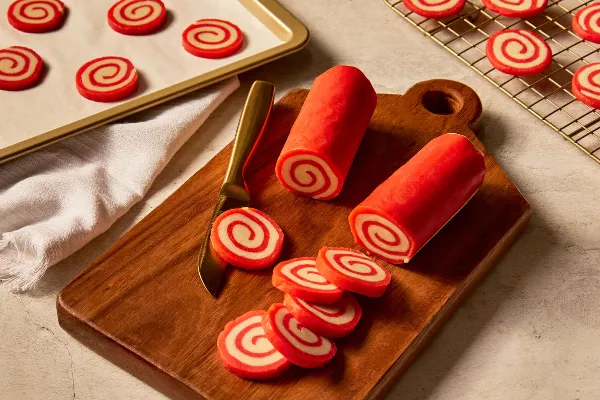 Deux rouleaux de pâte à biscuits en spirale aux couleurs rouge et blanche, posés sur une planche à découper en bois avec plusieurs tranches coupées, et d'autres tranches sur une plaque de cuisson, ainsi que des biscuits cuits refroidissant sur une grille en métal