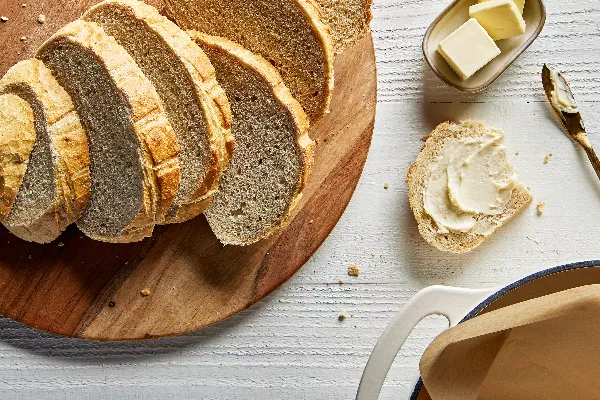 Un pain de levain sucré coupé en tranches sur une planche à découper en bois, avec une tranche beurrée, présenté avec un plat de beurre et un couteau à beurre.